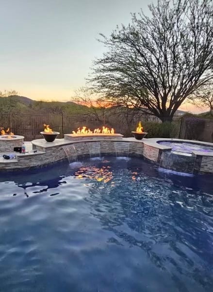 Custom pool with a stacked-stone spa, waterfall spillways, and fire features at dusk in Tucson by Sorce Signature Outdoors